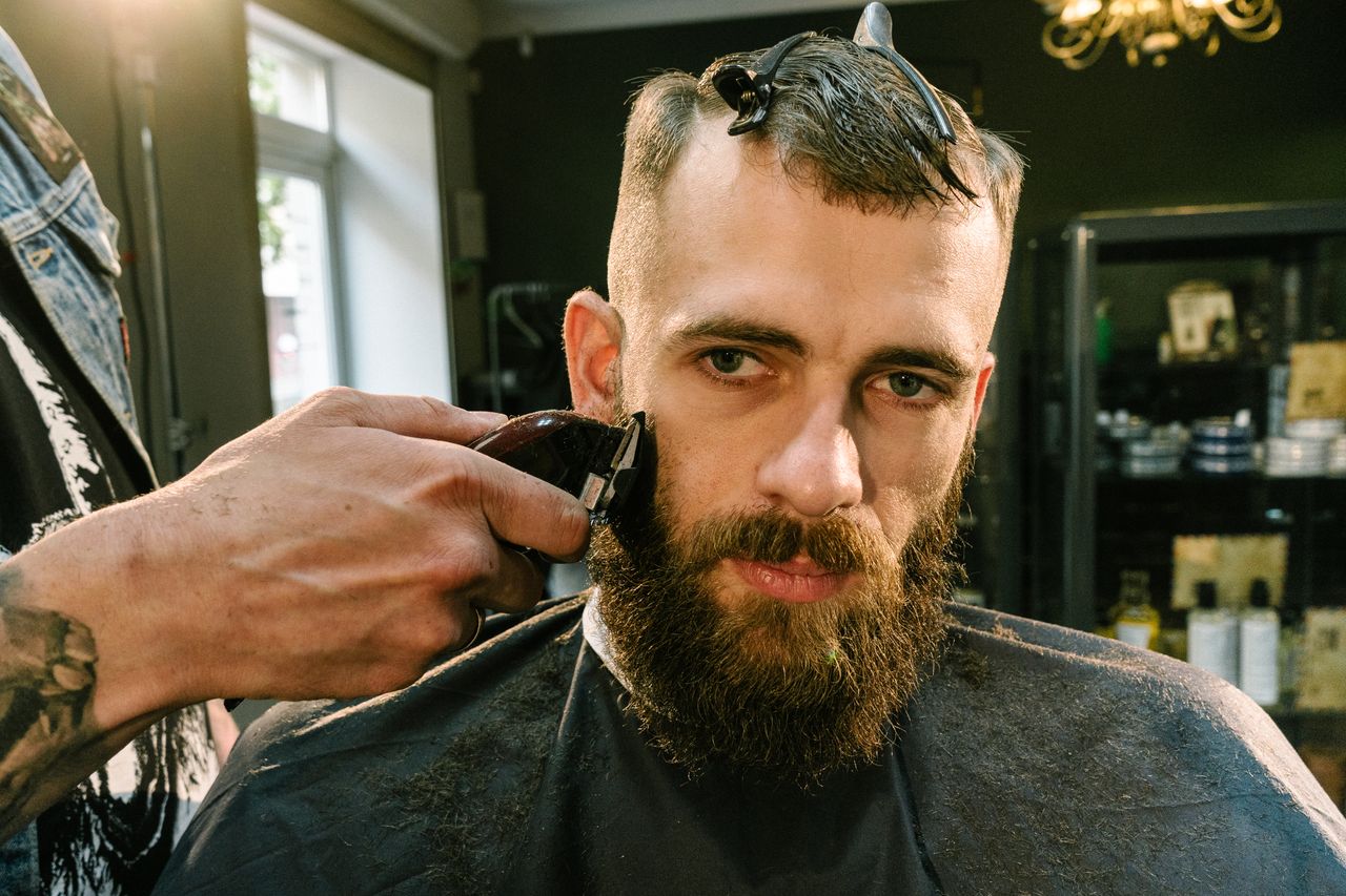 contact-img Close-up of a barber trimming a bearded man's facial hair in a stylish barber shop.