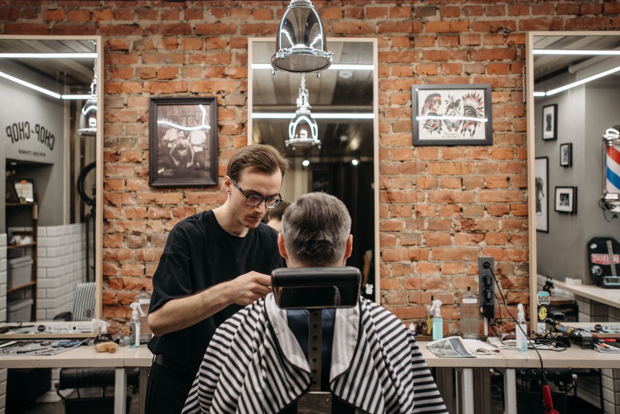 A barber cuts a customer's hair in a stylish, industrial-themed barbershop.
